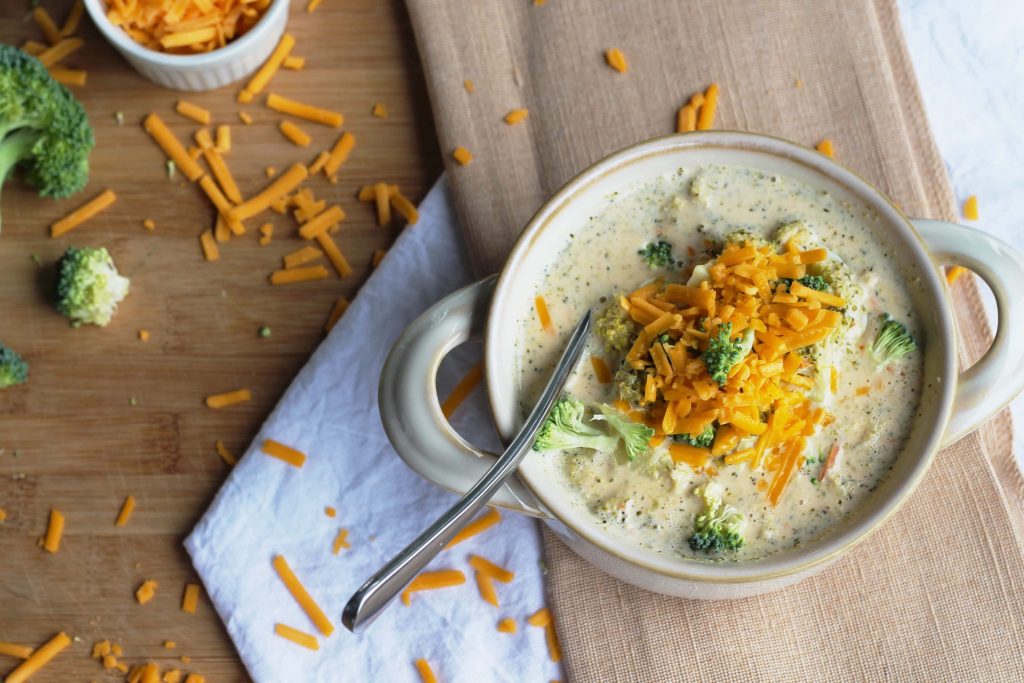 slow cooked broccoli cheese soup topped with extra broccoli and shredded sharp cheddar cheese in a beige bowl on top of a table cloth with a side dish of cheese.