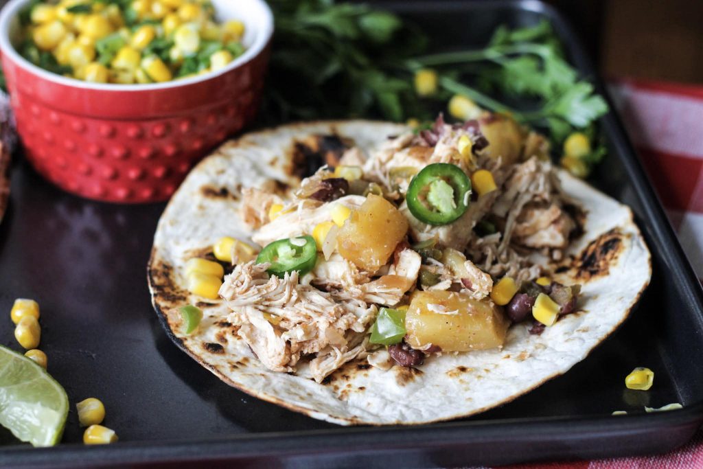 chicken, and diced pineapples, with jelopanos,and corn on a crisp flour tortilla, on a one pan baking sheet with lies and a side of corn and green peppers in a red bowl.