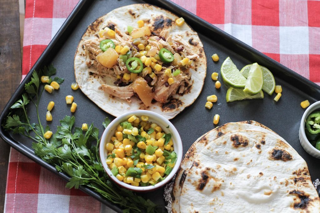 chicken, and diced pineapples, with jelopanos,and corn on a crisp flour tortilla, on a one pan baking sheet with lies and a side of corn and green peppers in a red bowl, with a side of cilantro.