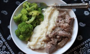 slowed cooked and seasoned beef stroganoff, served with mashed potatoes, and steamed broccoli on a white plate with a silver fork.