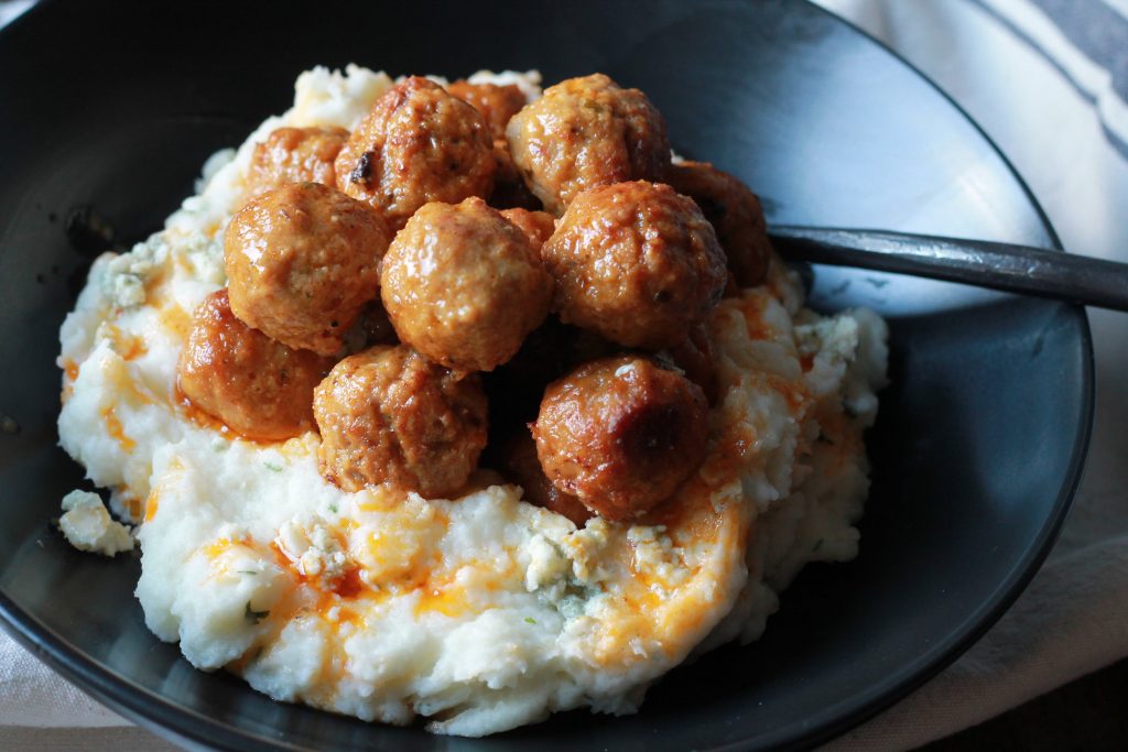 slow cooked meatballs topped with spicy buffalo sauce served on top of mashed potatoes in a black bowl.