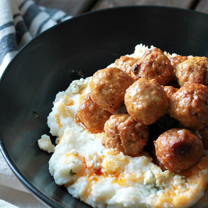 slow cooked meatballs topped with spicy buffalo sauce served on top of mashed potatoes in a black bowl.