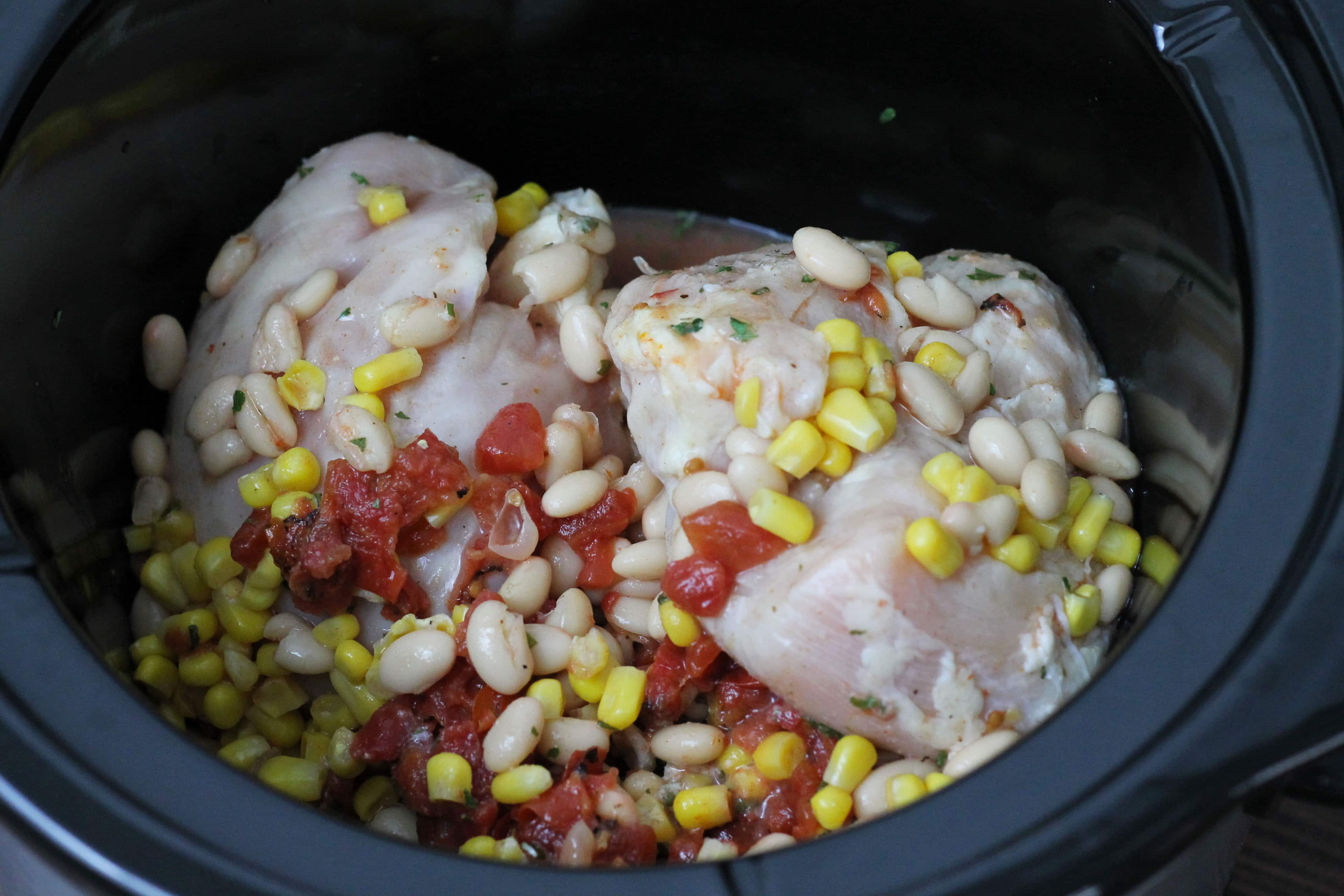 uncooked chicken with corn diced tomatoes beans in a black crock pot.