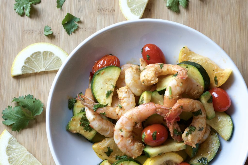 cooked shrimp with lemon slices, petit tomoates, with sliced zucchini topped with cilantro in a light blue bowl on a wooden table with a slice of lemon and pieces of cilantro on the table.