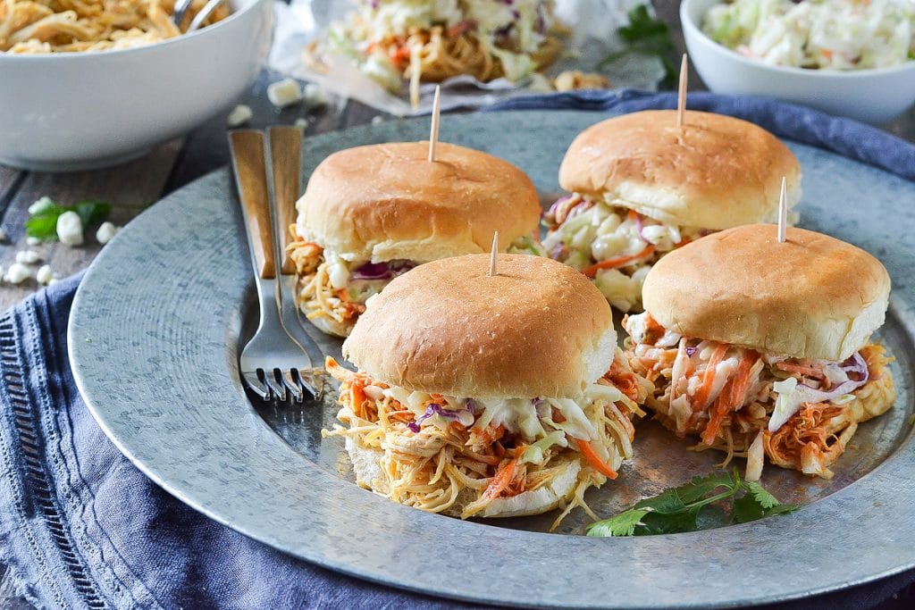 4 buffalo chicken sliders with cole slaw on a round metal plate with forks and blue napkin