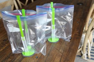 two green Jokari Hands-Free Bag Holders, holding up clear zip lock gallon bags on a wooden table 