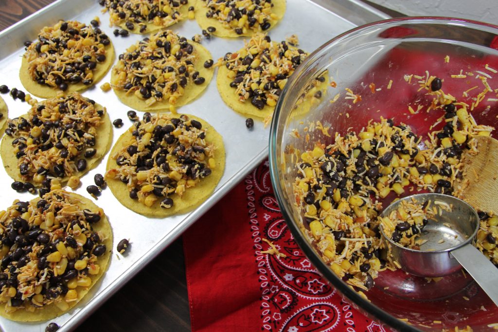 black beans, shredded cheese and corn mixed in a clear bowel being served onto seven crispy tostadas on a baking sheet.