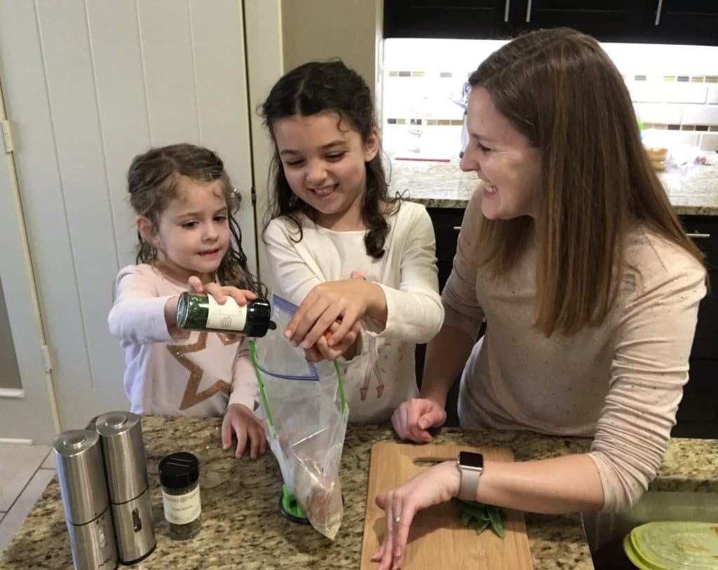 A Mom with her two daughters in the kitchen cooking, and laughing while preparing dinner .