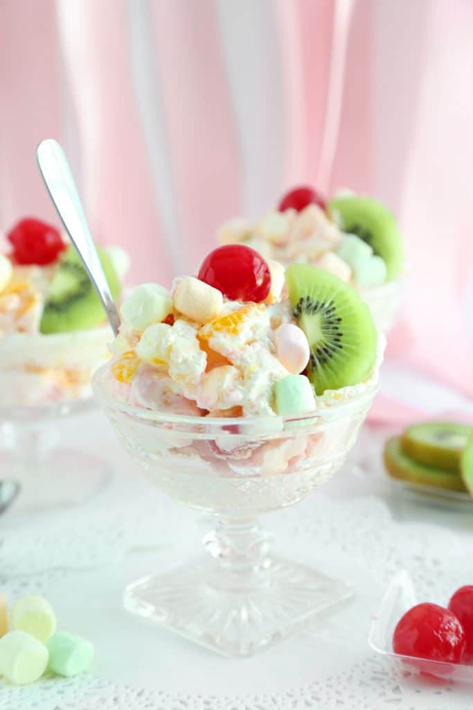 Fresh fruit whipped cream served with colorful marshmallows a kiwi slice and topped with a red cherry all served in an ice cream dish. 