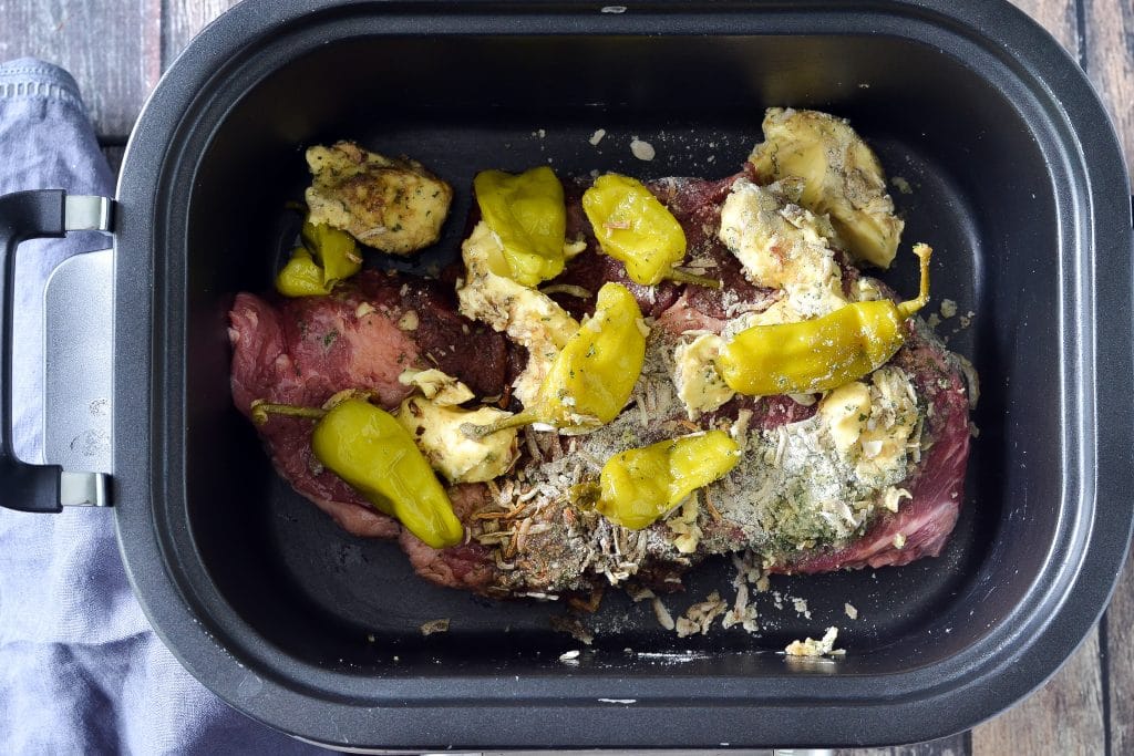 Uncooked pot roast, olive oil, ranch mix, onion soup mix, pepperoni peppers, and chunks of butter all mixed together and placed in a crock pot to be cooked.