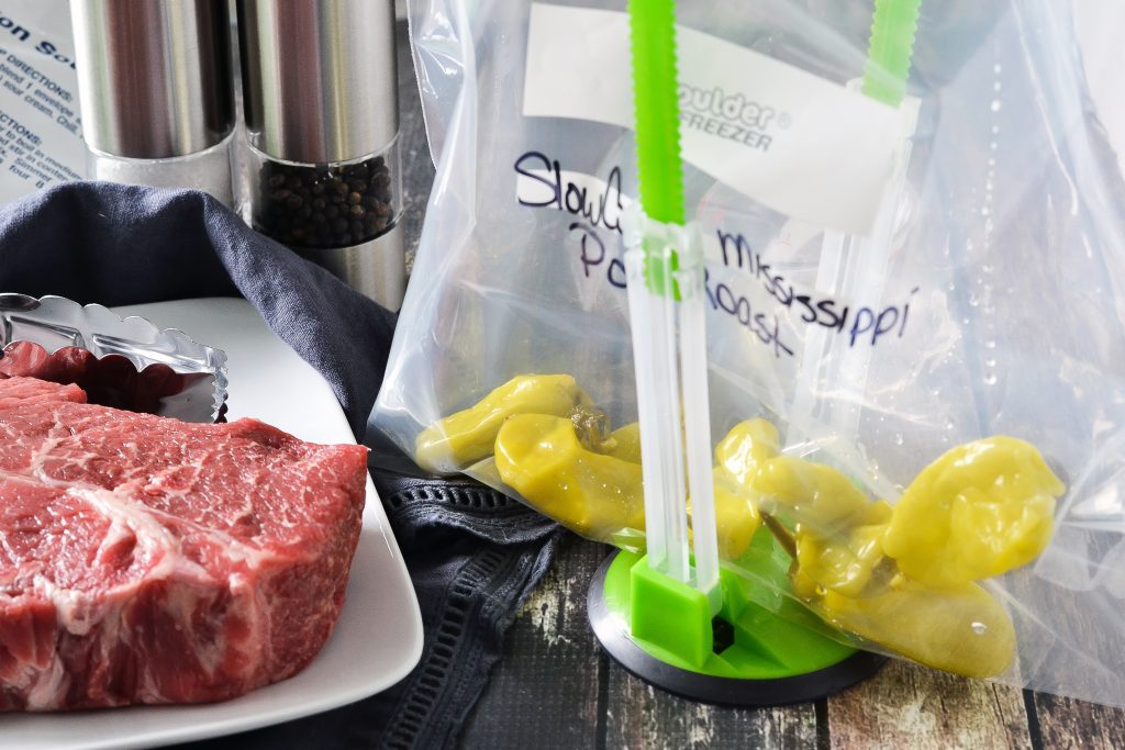 A labeled plastic bag with pepperoni peppers inside, being held by green baggie holder, on the counter next to an uncooked pot roast on a plate.