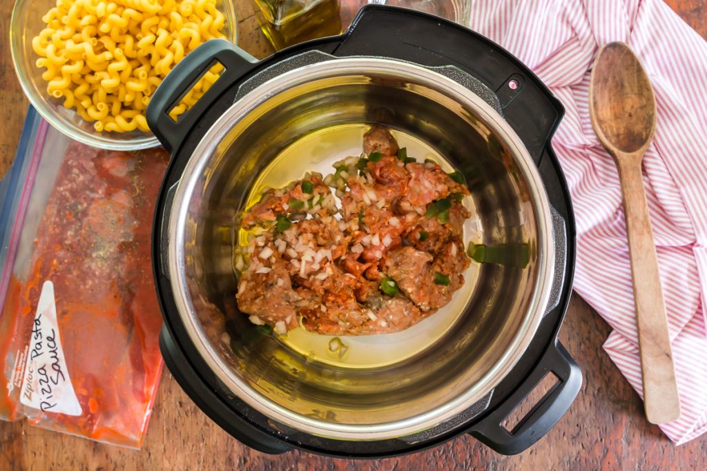 Instant pot with ground beef browning and a side of dried pasta and wood spoon