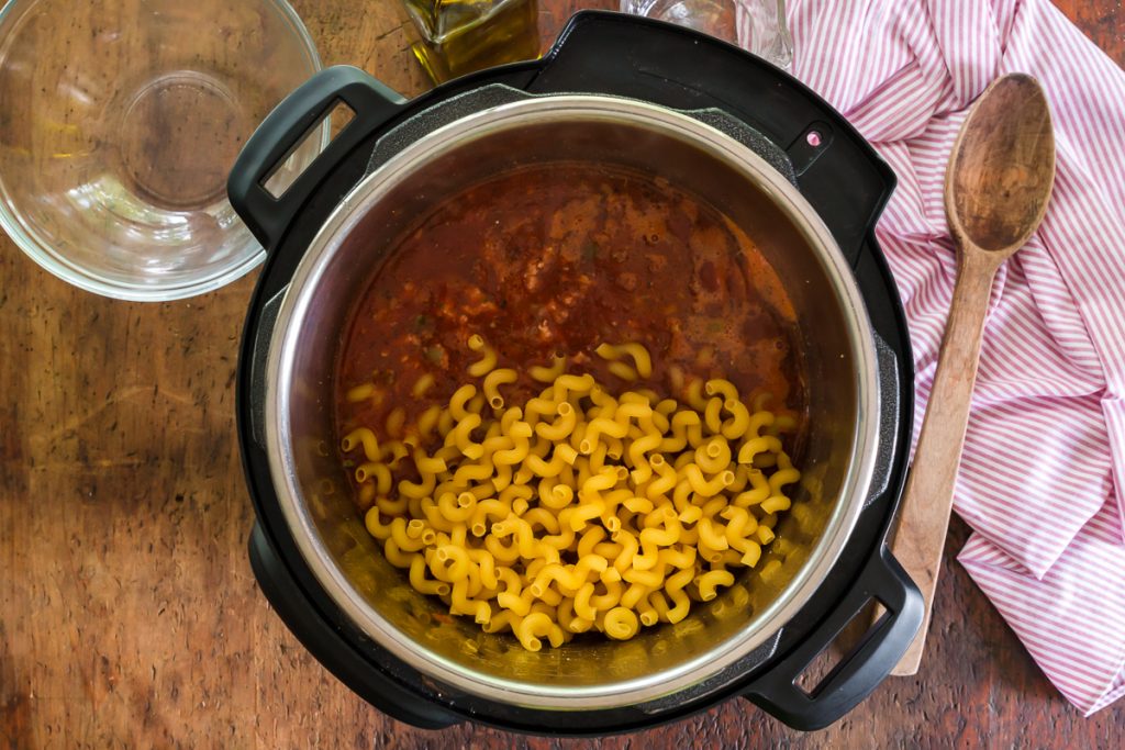 instant pot fille with dried pasta, ground beef and tomato sauce, empty clear bowl and wood spoon on sides of pot